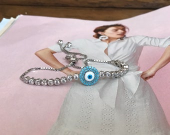 Puede incluir: Una pulsera de plata con un dije de ojo turco azul rodeado de piedras de circonita cúbica transparentes. La pulsera tiene un cierre deslizante ajustable.