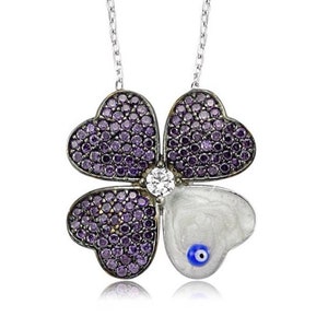 Może przedstawiać: Srebrny naszyjnik z zawieszką w kształcie koniczyny czteroliściennej. Koniczyna jest wykonana ze srebra i ma w każdym listku osadzone fioletowe kamienie. W środku koniczyny znajduje się biały kamień i niebieski amulet.