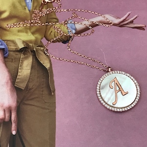 Peut inclure: Un collier en chaîne dorée avec un pendentif rond orné d'une incrustation de nacre blanche et d'une lettre dorée 'A' entourée de strass transparents.
