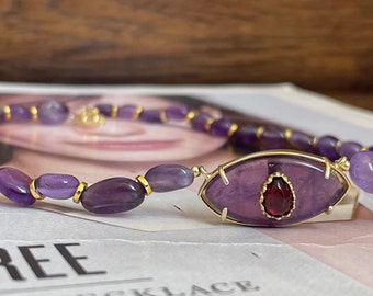 Könnte beinhalten: Eine Halskette aus violetten Amethyst-Edelsteinen mit einem goldenen, drahtgewickelten Anhänger, der einen größeren Amethyststein mit einem roten Granat-Akzent zeigt. Die Halskette liegt auf einem Magazin mit dem aufgedruckten Wort "THREE".