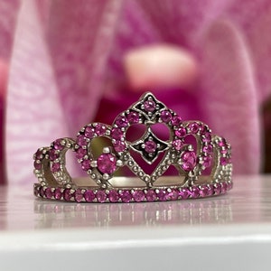 May include: A silver crown ring with pink gemstones. The crown has a heart-shaped design and is set with small, sparkling stones.
