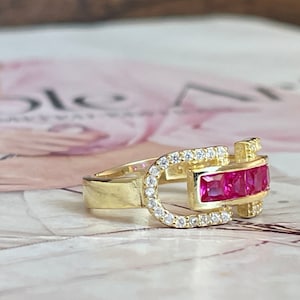 May include: A gold ring with a red stone and clear stones set in a buckle design.
