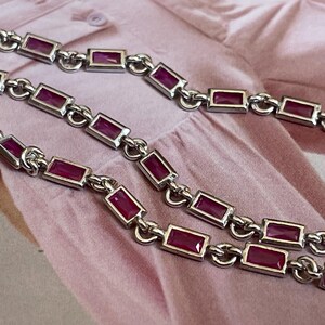May include: Close-up of a silver necklace with rectangular, deep red gemstones. The necklace is composed of connected links, each holding a gemstone. The background is a pink fabric.