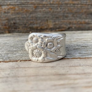 Puede incluir: Un anillo de plata con un diseño floral. El anillo está hecho de una cuchara reutilizada y presenta tres flores con tallas detalladas.