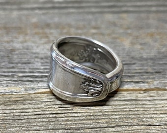 May include: A silver spoon ring featuring a floral design. The ring is crafted from a repurposed spoon and has a distinctive, rustic appearance.