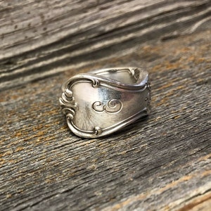 Könnte beinhalten: Ein silberner Löffelring mit einem dekorativen Design und den eingravierten Initialen "C.G." auf der Vorderseite.