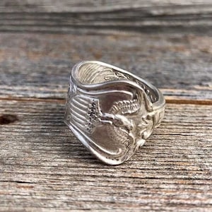 May include: A silver ring made from a spoon with an eagle design. The ring is on a wooden surface.