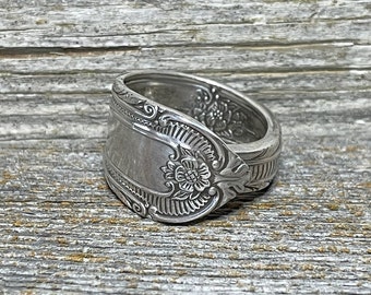 May include: A silver spoon ring featuring an ornate floral design. The ring is crafted from a vintage spoon and has a unique, handcrafted appearance.