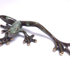 May include: A ceramic frog figurine with a glossy finish, featuring a dark brown body with light green mottled spots and metallic gold-toned accents on the tips of its splayed toes and fingers, captured in a low-profile, crawling pose.