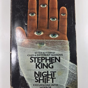 May include: A paperback book titled Night Shift by Stephen King with a black cover featuring a brown hand wrapped in white gauze bandages. The hand has multiple human eyes embedded in the skin. Text includes By the author of Cujo & Different Seasons.