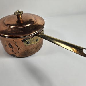 May include: A vintage copper saucepan with a brass handle and lid. The saucepan has a worn, aged look and is perfect for cooking on the stovetop.