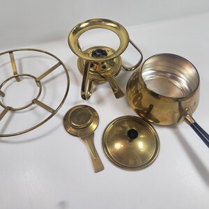 May include: A collection of vintage brass serving pieces, including a chafing dish, a warming stand, a lid, and a ladle. The chafing dish has a black handle. The pieces are arranged on a white surface.