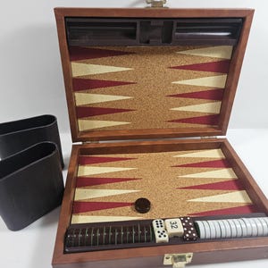 May include: A brown wooden backgammon set with a corkboard playing surface featuring red and beige triangles. Includes dice, checkers, and two black leather-like dice cups. The set is open, displaying the game pieces.