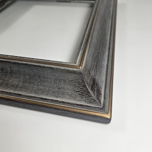 May include: Close-up of a rectangular picture frame with a distressed, weathered appearance. The frame is primarily dark gray with white and gold accents. The frame's corner is visible, showcasing its construction.