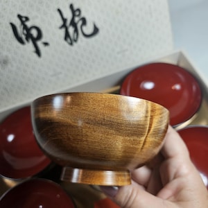 May include: A wooden bowl with a natural wood grain finish, held in a hand. The bowl is part of a set that includes several red lacquered bowls. The box has Japanese characters.