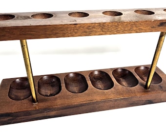 May include: A dark brown wooden stand featuring two horizontal rectangular blocks connected by two thin brass-colored metal rods. The top block contains six circular indentations, while the bottom block features six oval-shaped carved wells.