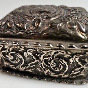May include: A small, antique silver box with a hinged lid. The box features ornate floral and scrollwork designs on the lid and sides. The edges have a scalloped trim. The box is likely made of sterling silver.