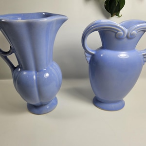 May include: Two matching light blue ceramic vases. The vase on the left has a handle and a bulbous body with vertical ridges. The vase on the right has two curved handles and a smooth, rounded body. Both vases have a pedestal base.