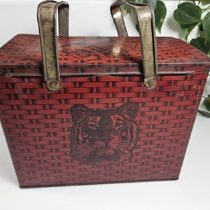 May include: A red and black vintage tin lunchbox with a tiger design and two metal handles. The lunchbox has a woven pattern.