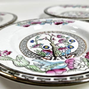 May include: Three white ceramic saucers featuring a central floral motif with pink, blue, and green colors. Each saucer has a black and white geometric border pattern along the rim, accented with a thin gold line, and a visible crackle glaze finish.