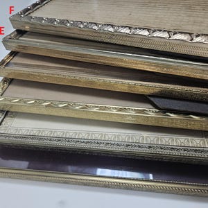 May include: A stack of six antique picture frames with ornate gold and silver-toned borders. The frames vary in style, with some featuring intricate patterns and others a more simple design. The frames are empty, ready for photos or artwork.