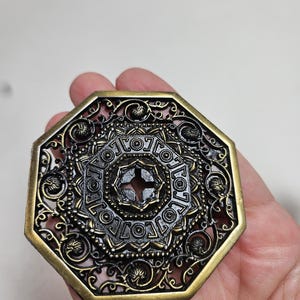 May include: Ornate, octagonal metal object with a detailed, filigree design. The object has a dark bronze center and a gold-colored border. The intricate pattern includes floral and geometric elements, creating a vintage or antique aesthetic.