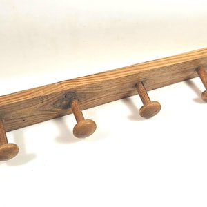 May include: A long, rectangular wooden wall rack featuring a natural brown wood grain finish. The rack is equipped with seven evenly spaced, rounded wooden pegs extending outward, and includes two small metal nails protruding from the board.