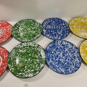 May include: A collection of colorful enamelware plates. The plates are in red, green, blue, and yellow, each with a white swirl pattern. The plates have a black rim and are arranged on a white surface.