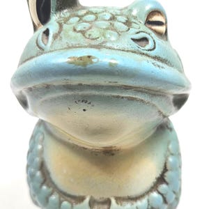 May include: A ceramic frog figurine featuring a light blue glaze with dark brown accents in the recessed areas. The frog has a textured head, prominent eyes, and a wide mouth. The body shows a rounded shape with visible molded details and markings.