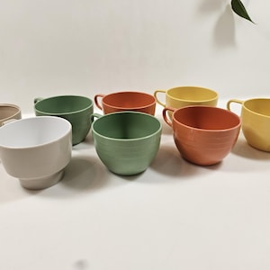 May include: A collection of eight colorful ceramic cups in various shades, including beige, green, red, yellow, and peach. Each cup has a handle and a smooth, glossy finish. The cups are arranged on a white surface.