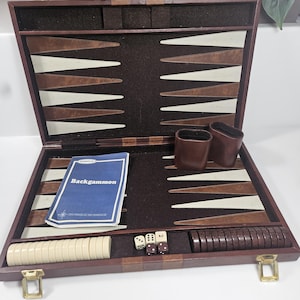 May include: Open backgammon set with a dark brown wooden case, featuring a felt playing surface with brown, white, and tan triangles. Includes dice, game pieces, dice cups, and a rule book titled "Backgammon".