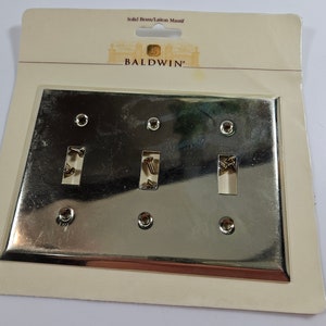 May include: A gold-colored metal switch plate with three switch openings and six screw holes. The plate is packaged in a white cardboard backing with the text "Solid Brass/Laiton Massif" and "BALDWIN".