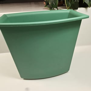 May include: A green, trapezoidal-shaped planter with a wide, flat rim. The planter has a smooth, matte finish and is designed to hold plants. The color is a muted, earthy green, suitable for various home decor styles.