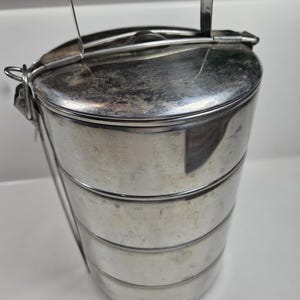 May include: A vintage, stainless steel tiffin carrier with four stacked compartments. The carrier has a handle for easy transport and a secure latching system. The metal has a brushed finish and shows signs of wear, suggesting its age and use.