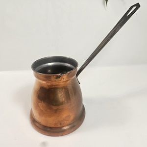 May include: A copper-colored Turkish coffee pot with a long, dark handle. The pot has a rounded base and a cylindrical body, with a dark interior. The handle extends from the side and curves upward. The pot is likely made of copper.