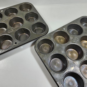 May include: Two black metal muffin tins. The tin on the left has a smooth surface and the tin on the right has a decorative starburst pattern.