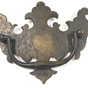 May include: A dark bronze-toned metal drawer pull featuring a decorative, ornate backplate with symmetrical scrollwork and a curved, swinging handle attached to two circular mounting points. The surface shows a weathered, aged patina finish.