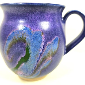 May include: A rounded ceramic mug featuring a glossy glaze in shades of deep purple and royal blue. The surface displays a swirling, abstract pattern with accents of bright blue and a small patch of dark green, finished with a curved handle.
