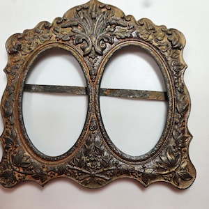 May include: Ornate, antique-style double oval picture frame in bronze and gold tones. The frame features detailed floral and leaf designs, with two oval openings and a horizontal bar across each opening.