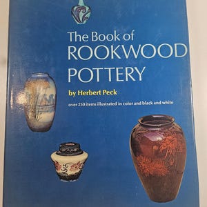 May include: A blue book cover with the title "The Book of Rookwood Pottery" and author Herbert Peck. The cover features illustrations of Rookwood pottery vases in various colors and designs.