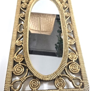May include: A rectangular wall mirror featuring an oval center glass panel surrounded by a gold-toned frame. The frame displays a textured, ribbed border and decorative scrollwork patterns in the corners and along the sides of the oval.