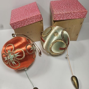 May include: Two vintage Christmas ornaments with matching boxes. One ornament is orange with gold trim and mint green accents. The other is pale green with gold trim and red ribbon. Each has a gold-toned teardrop-shaped pendant.