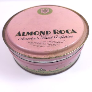 May include: A round pink metal tin for Almond Roca candy. The lid features the text Almond Roca, America's Finest Confection, and a list of ingredients including butter, milk chocolate, sugar, almonds, coconut oil, salt, vanilla, and lecithin.