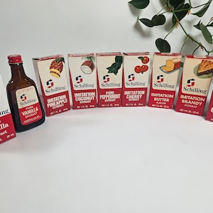 May include: A collection of eight small glass bottles of Schilling brand flavor extracts. The bottles are arranged in a row on a white surface. The flavors include: Pure Vanilla, Imitation Pineapple, Imitation Coconut, Pure Peppermint, Imitation Cherry, Imitation Butter, Imitation Brandy, and Pure Orange.