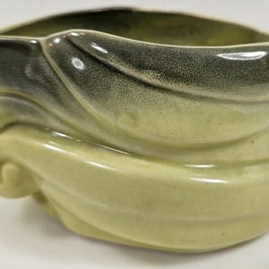 May include: A ceramic bowl featuring a glossy finish in shades of light olive green and dark charcoal gray. The exterior is molded with swirling, leaf-like organic shapes that create a layered, textured appearance around the entire vessel.