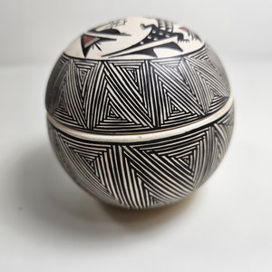 May include: A white ceramic round box with a black and white geometric pattern. The top of the box has a black and red lizard design.