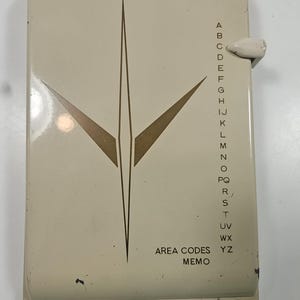 May include: Vintage cream-colored address book with a geometric design in gold. The book has a flip-up cover and a tabbed index for alphabetized entries. The text "AREA CODES MEMO" is printed on the front.