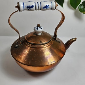 May include: A copper teapot with a white porcelain lid and handle. The handle has a blue and white floral design.