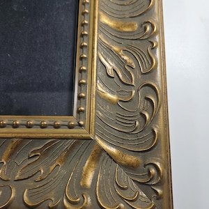 May include: Close-up of a gold-colored ornate picture frame with a detailed, carved design. The frame is made of wood and has a slightly distressed finish.