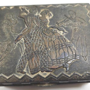 May include: A rectangular metal box with a dark, weathered finish. The lid features a raised, embossed scene of a couple dancing in the center, a figure playing a violin to the right, and a zigzag border pattern framing the entire top surface.